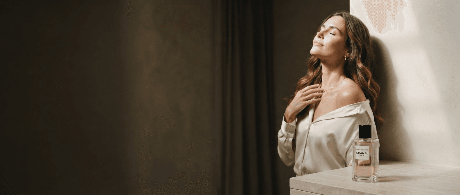 Mujer con blusa de seda junto a una ventana, con un frasco de perfume sobre la repisa